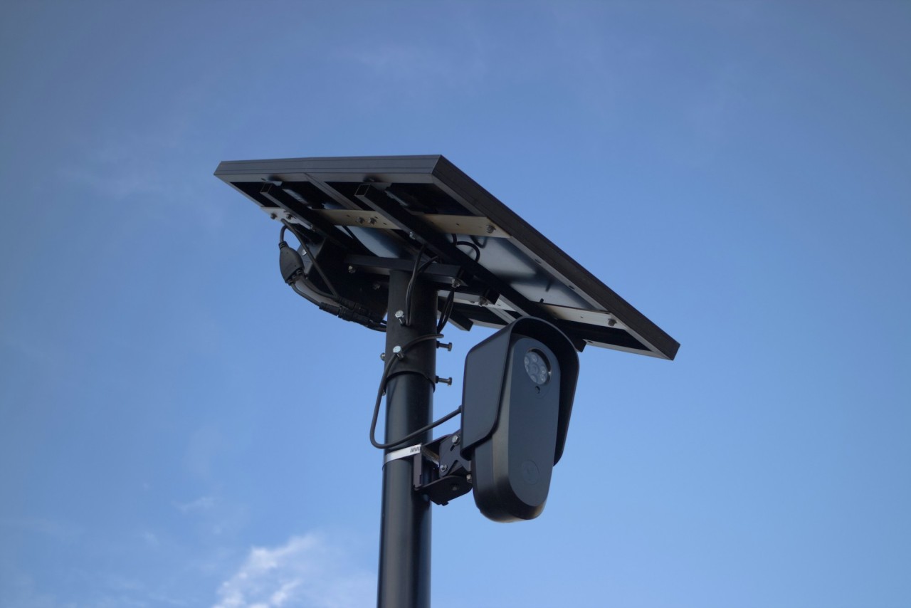 A solar-powered license plate recognition camera mounted on a black pole against a clear blue sky.
