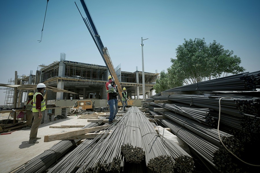 Workers in safety vests and helmets handle bundled steel rods with a crane at an active construction site where a multi-story concrete building framework is under development.