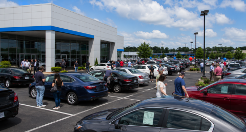 Busy car dealership lot with rows of parked vehicles and customers browsing cars outside a modern showroom on a busy sales day.